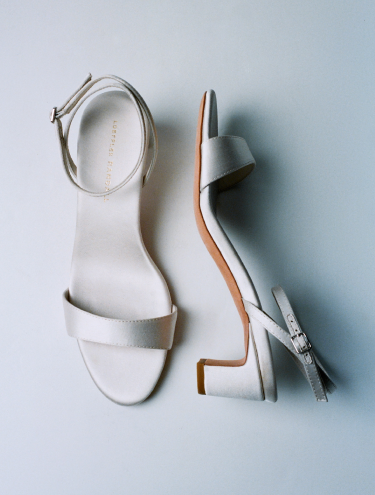 A pair of cream colored low heeled sandal with an ankle clasp one shoe is on its side on a grey background.