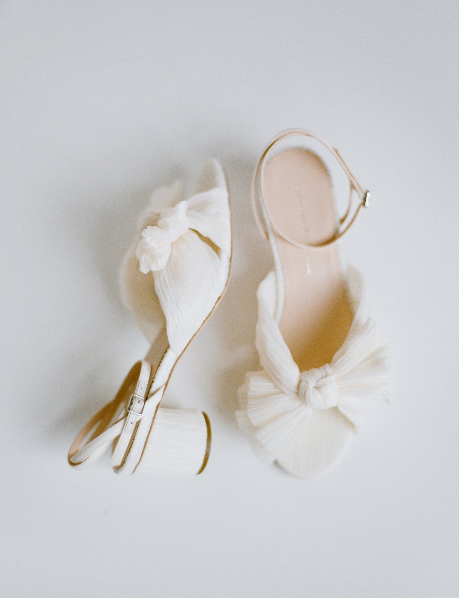 A pair of white heeled sandals with a pleated bow at the toes with one shoe on its side on a grey/white background.