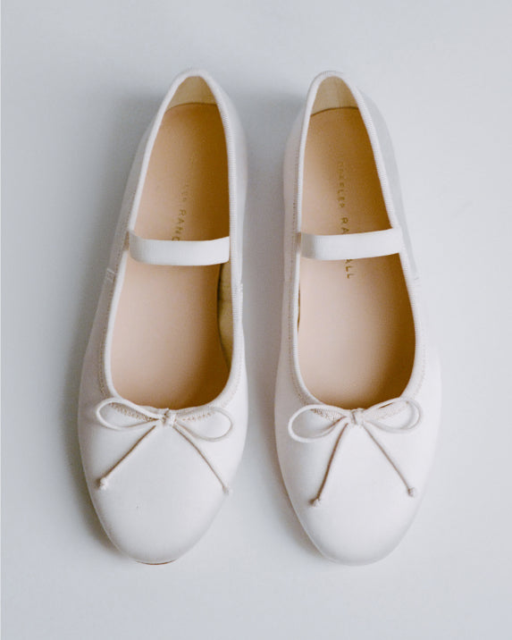 A pair of white flats with string bows at near the toes of the show on a grey background.