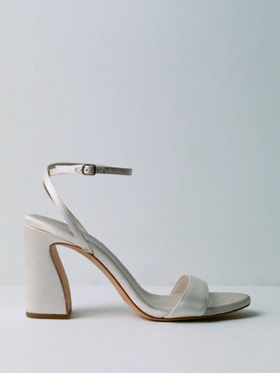 A side view of a white curved heeled sandal on a grey background.