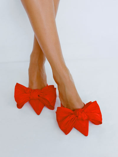 A model wears red bow heels standing in front of a grey background.