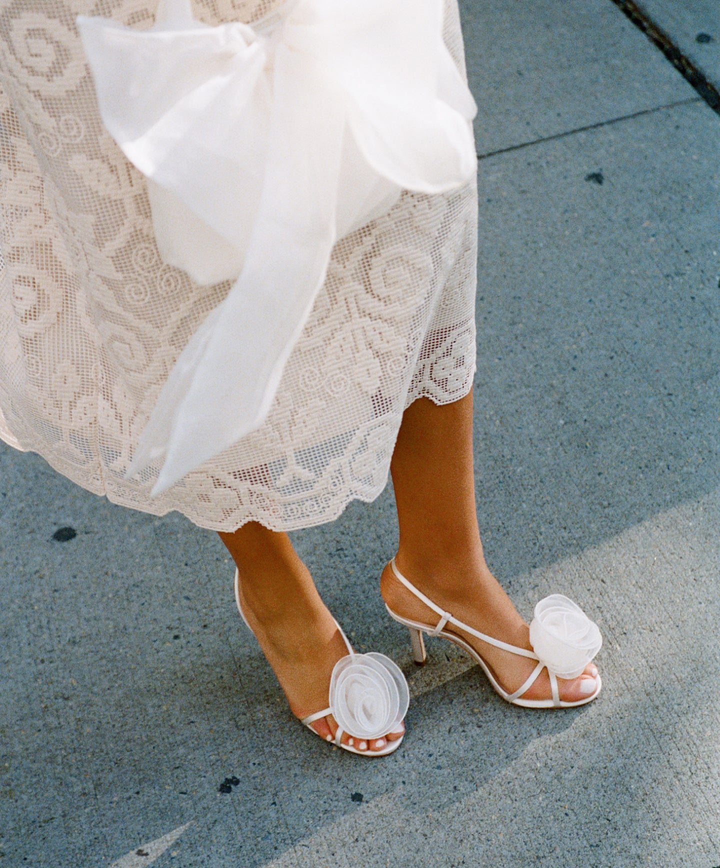 A model wears a white lace dress and white heels with a rose on the toes, holding a white bow clutch.
