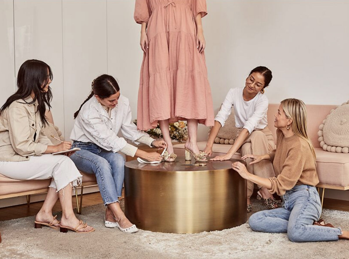 A model stands on a gold table wearing a pale pink dress and gold heeled sandals while four women sits on the ground and on couches surrounding her.