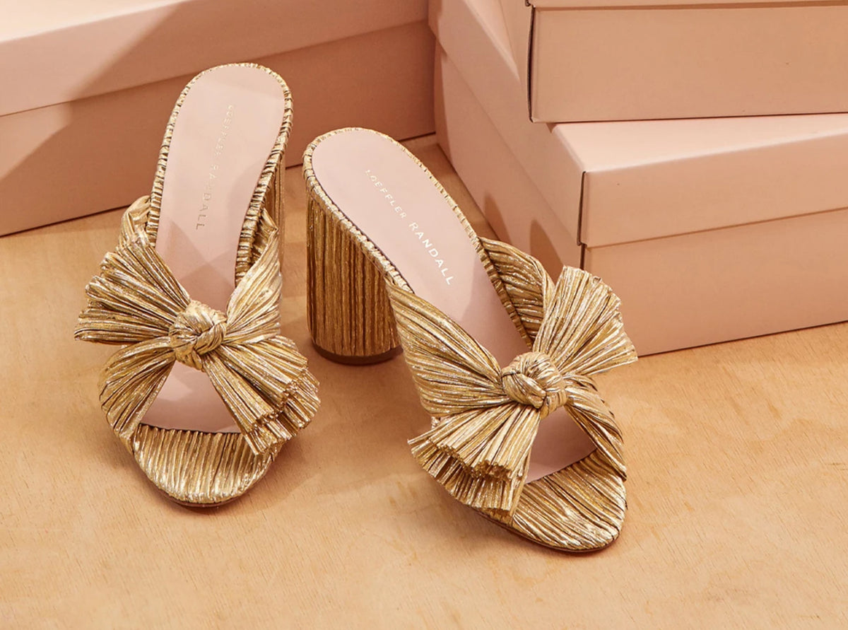 A pair of gold pleated bow heeled sandals in front of pink shoe boxes.