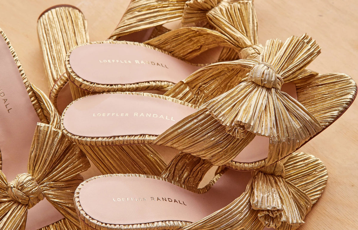 A close up of a pile of gold pleated heeled sandals on a wood background.
