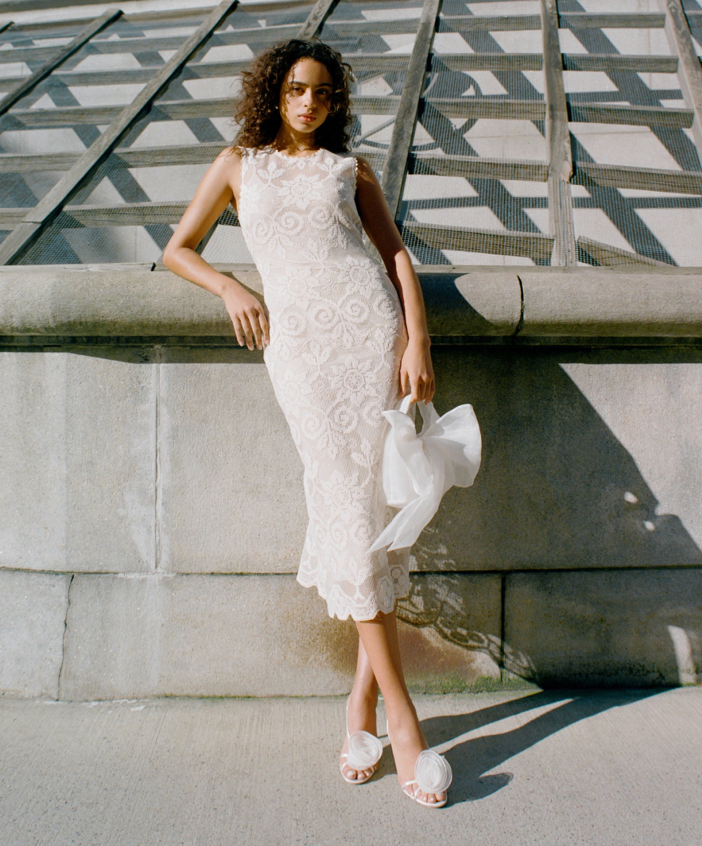 A model leans on a cement wall wearing a white lace dress and white heels with a rose on the toes, holding a white bow clutch.