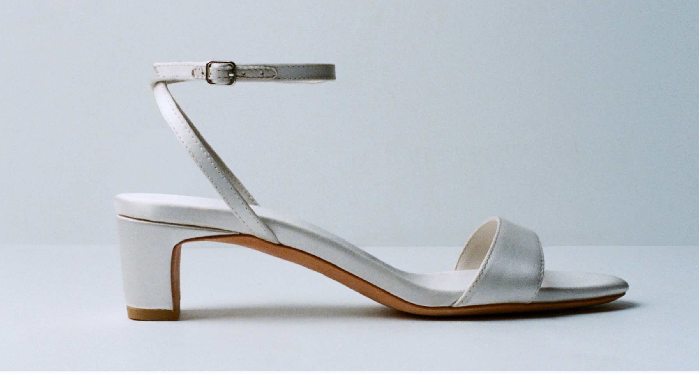 A cream colored low heeled sandal with an ankle clasp on a grey background.