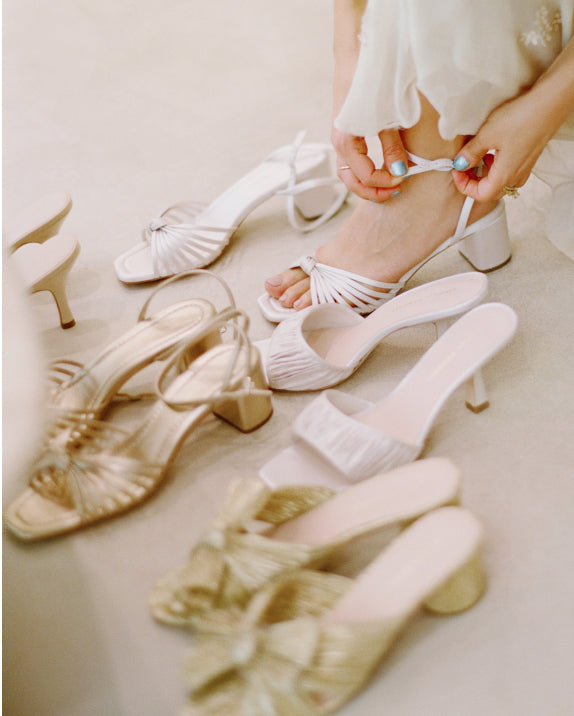 A model tries on a white heeled sandal with several other pairs of shoes on the floor beneath her.