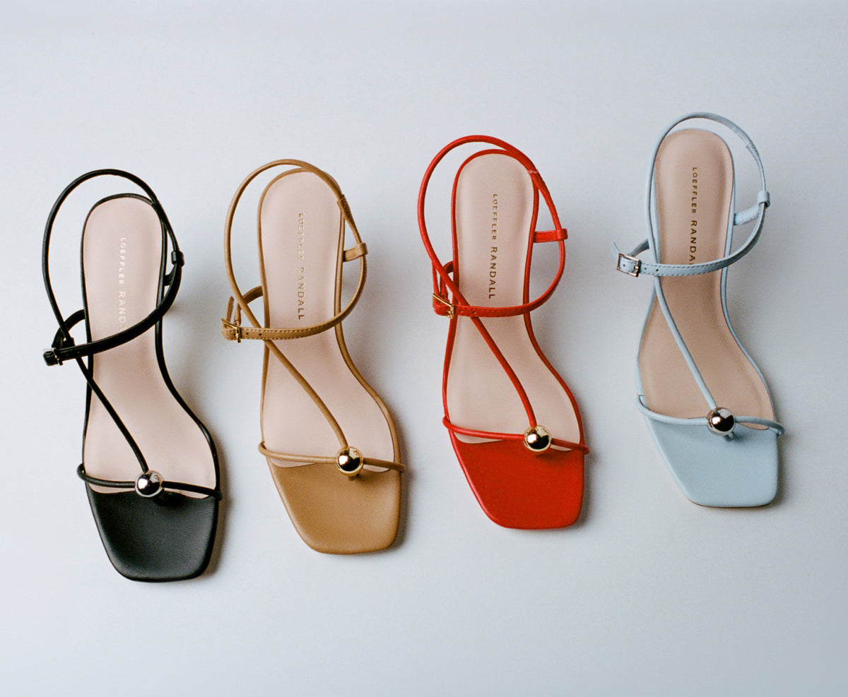 A cascade of different colored strappy heeled sandals with a gold accent near the toes on a grey background.
