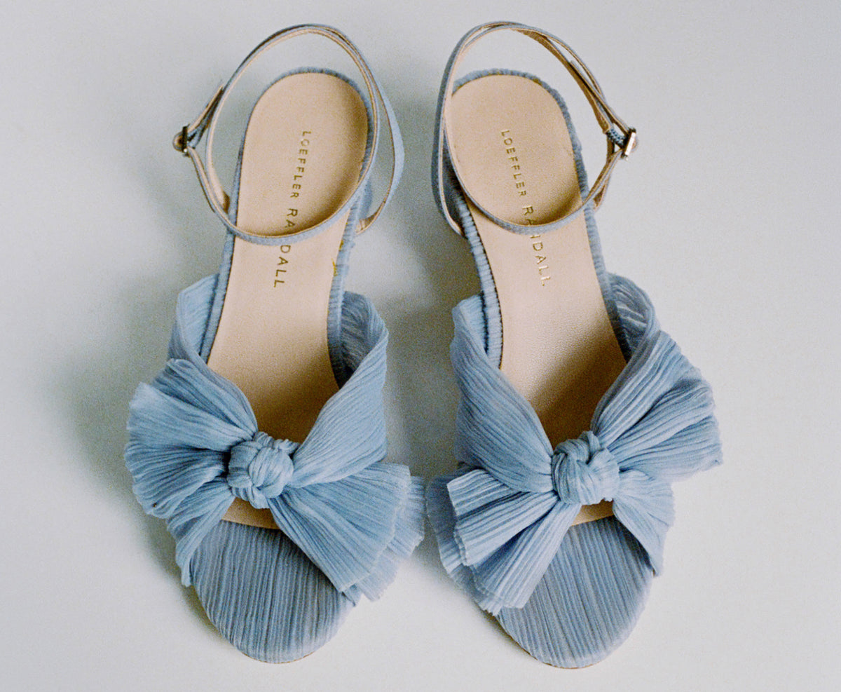 A pair of blue heeled sandals with pleated bows on the toe with a strap around the ankle on a grey background.