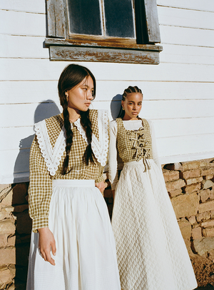 Two women wearing plaid tops and white skirts standing against a white and brick wall.