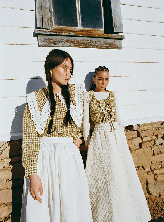 Two women wearing plaid tops and white skirts standing against a white and brick wall.