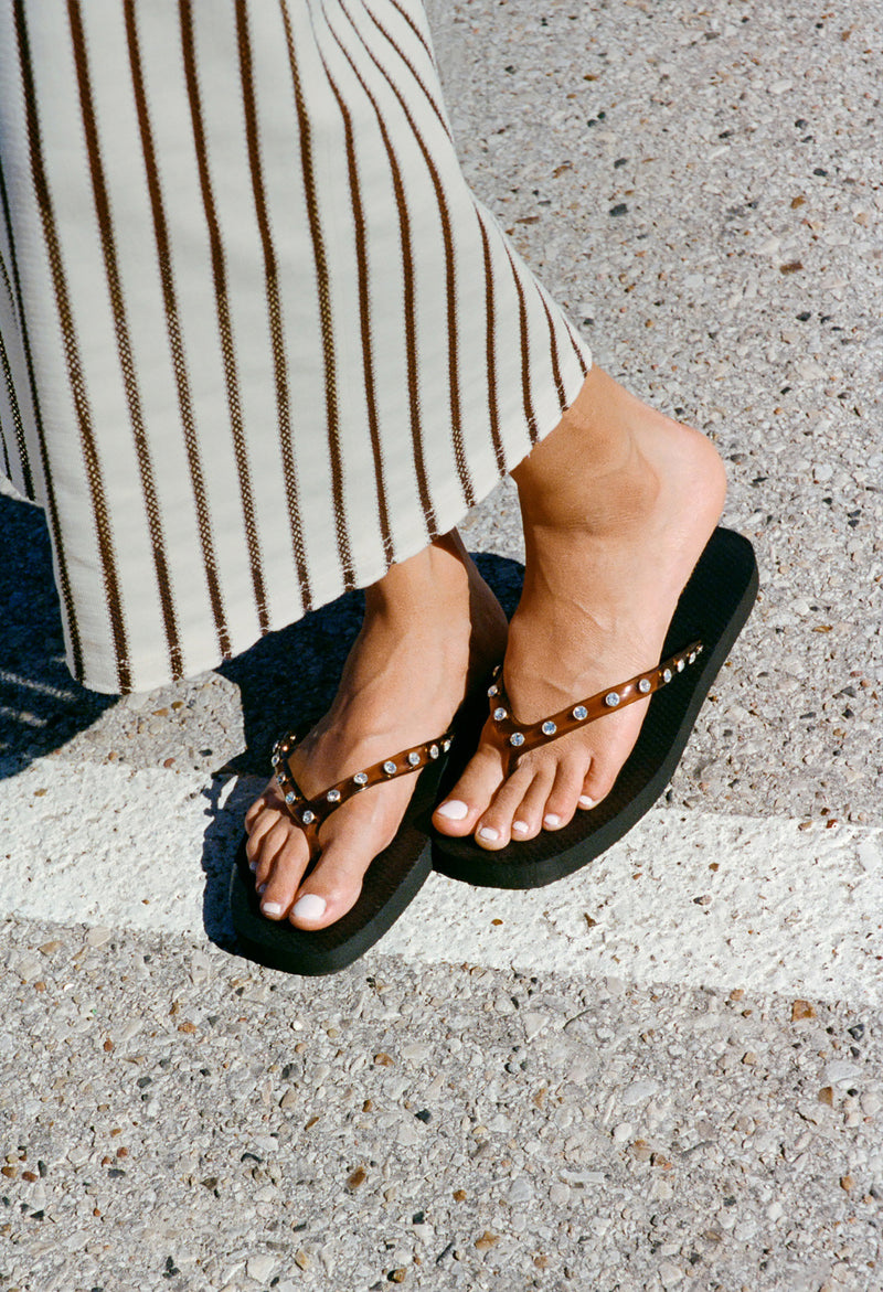 A model stands on a crosswalk wearing striped pants and brown flip flops with crystal details.