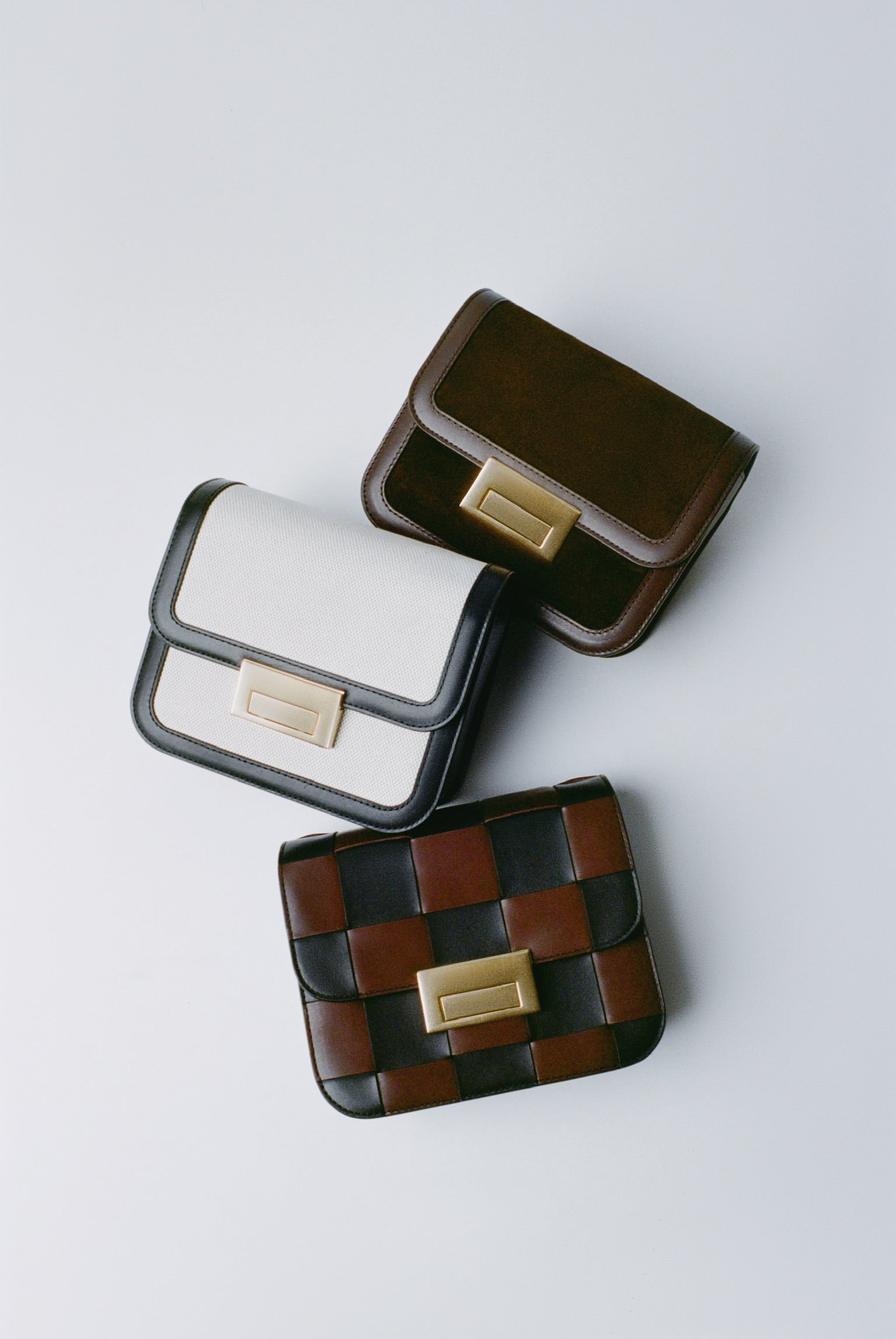 A cascade of three small handbags in different materials and colors with gold buckle accents on a light grey background.