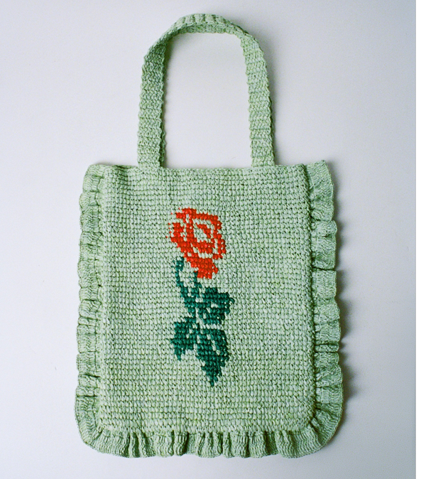 A mint green crochet tote with a red rose in the center on a grey background.