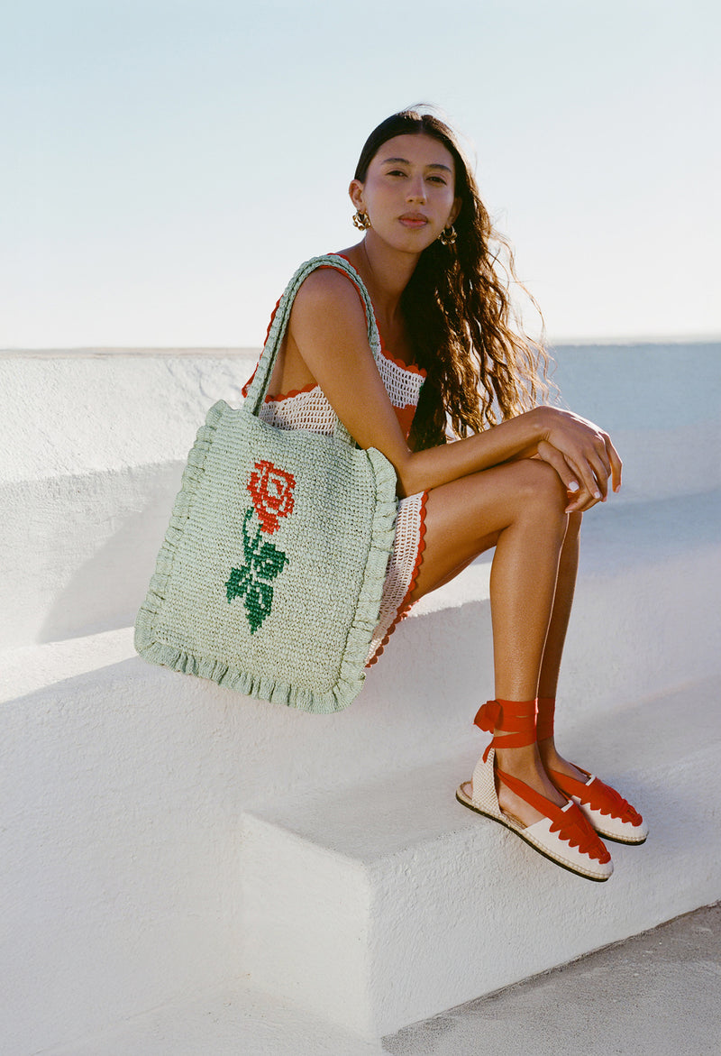 A model sits on a white stone wall wearing a white crochet dress, red lace up espadrilles, and a light green crochet tote with a rose on it.