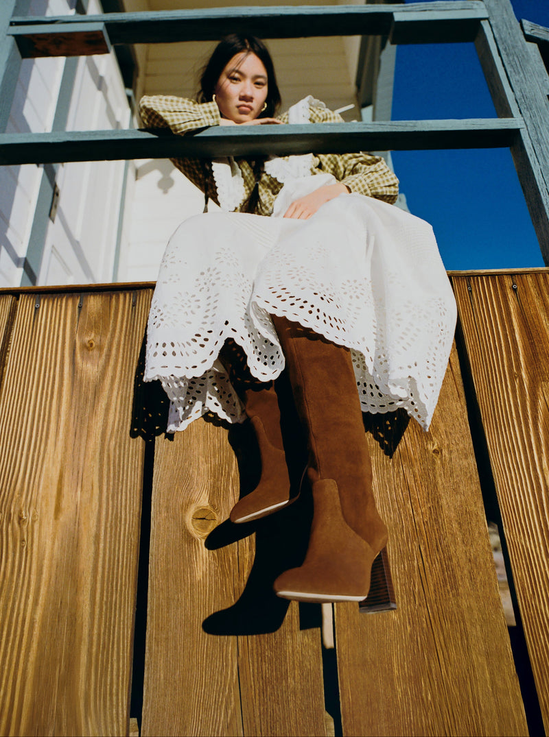 Woman sitting on a wooden railing wearing a white dress and brown suede tall boots.