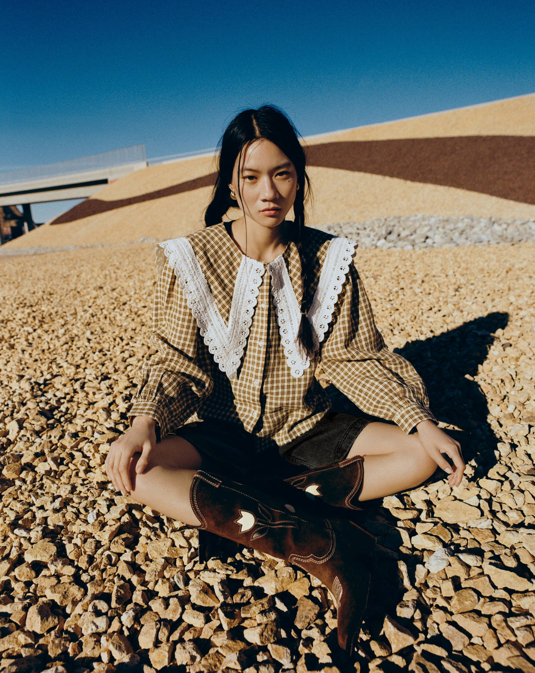 Woman sitting on a rocky surface wearing a checkered shirt and brown boots.