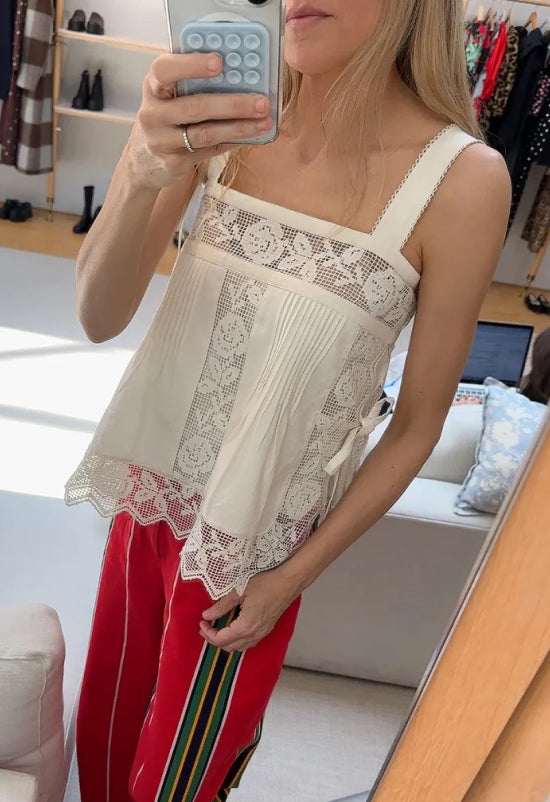 Jessie takes a mirror selfie in a store wearing a white tank top with lace embroidery designs and red pants.