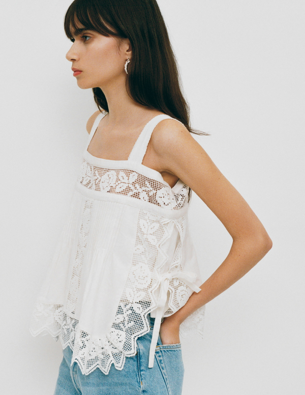 A model stands in front of a white background wearing a white tank top with lace embroidery details and light wash denim.