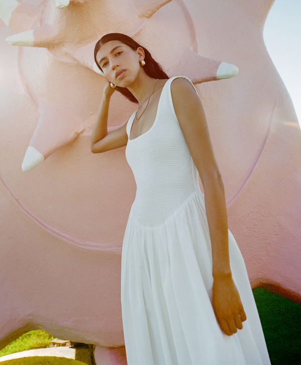 A model wears a white dress leaning against a pink statue.