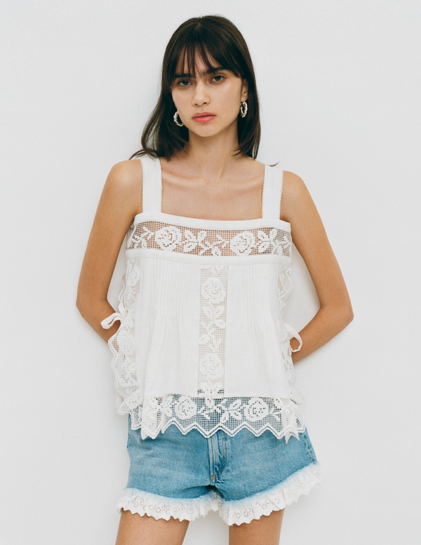 A model stands in front of a grey background wearing a white lace top and light denim shorts.