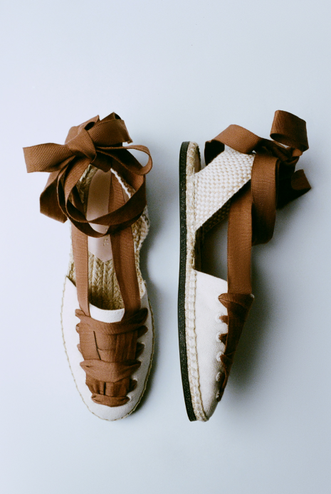 An overhead view of a pair of brown lace up espadrilles with one shoe on its side on a grey background.