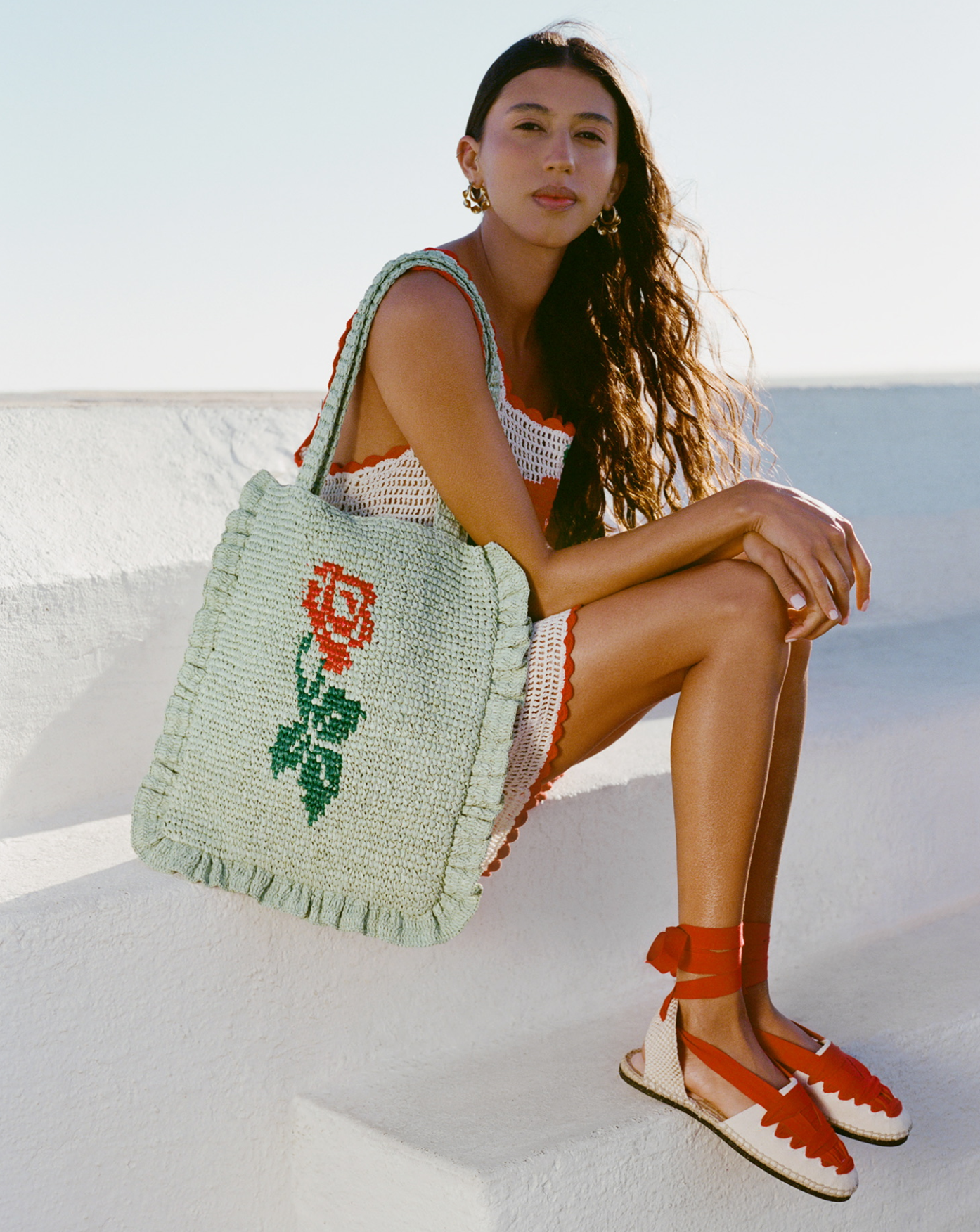 A model sits on a white stone wall wearing a white crochet dress, red lace up espadrilles, and a light green crochet tote with a rose on it.