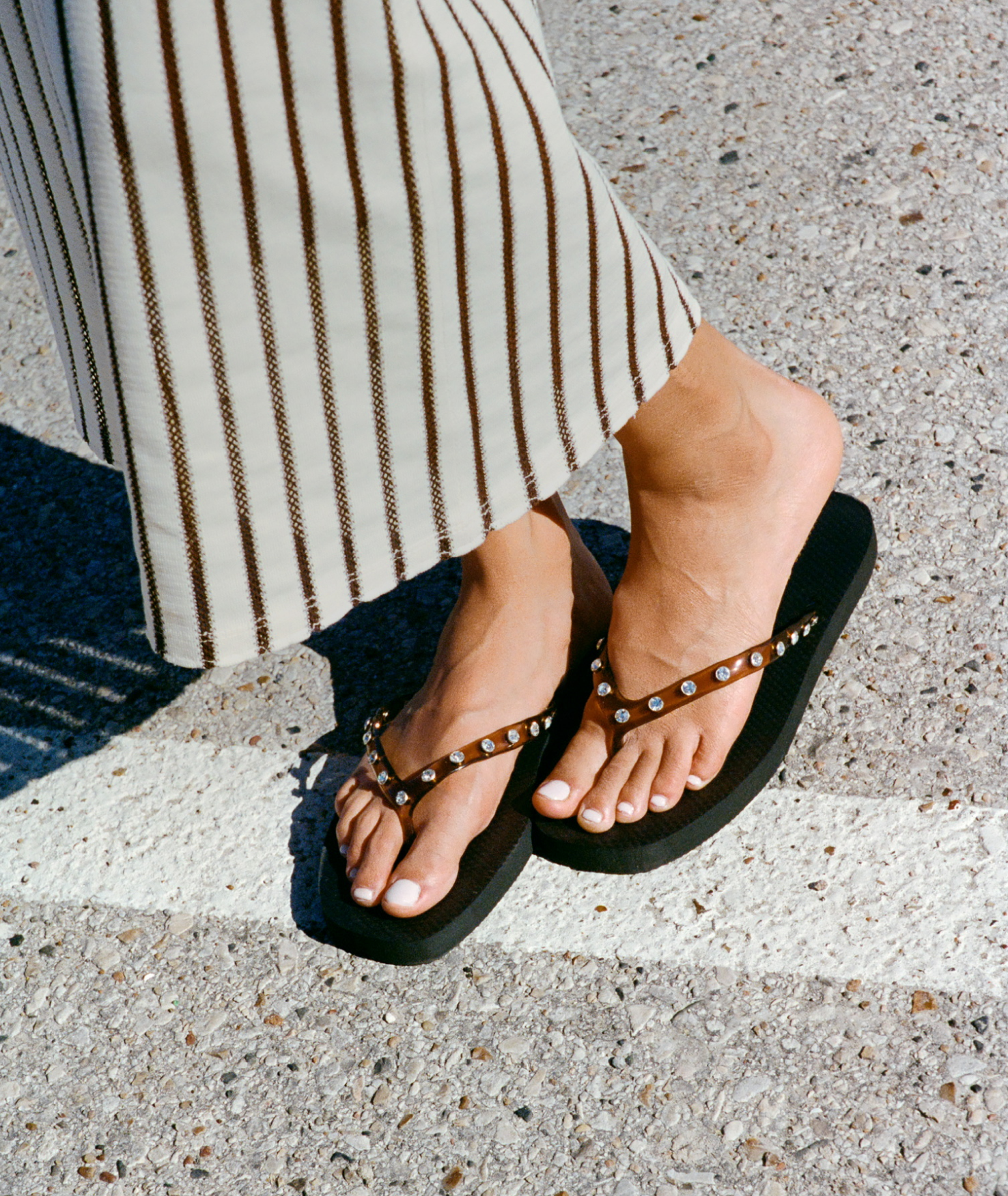 A model stands on a crosswalk wearing striped pants and brown flip flops with crystal details.