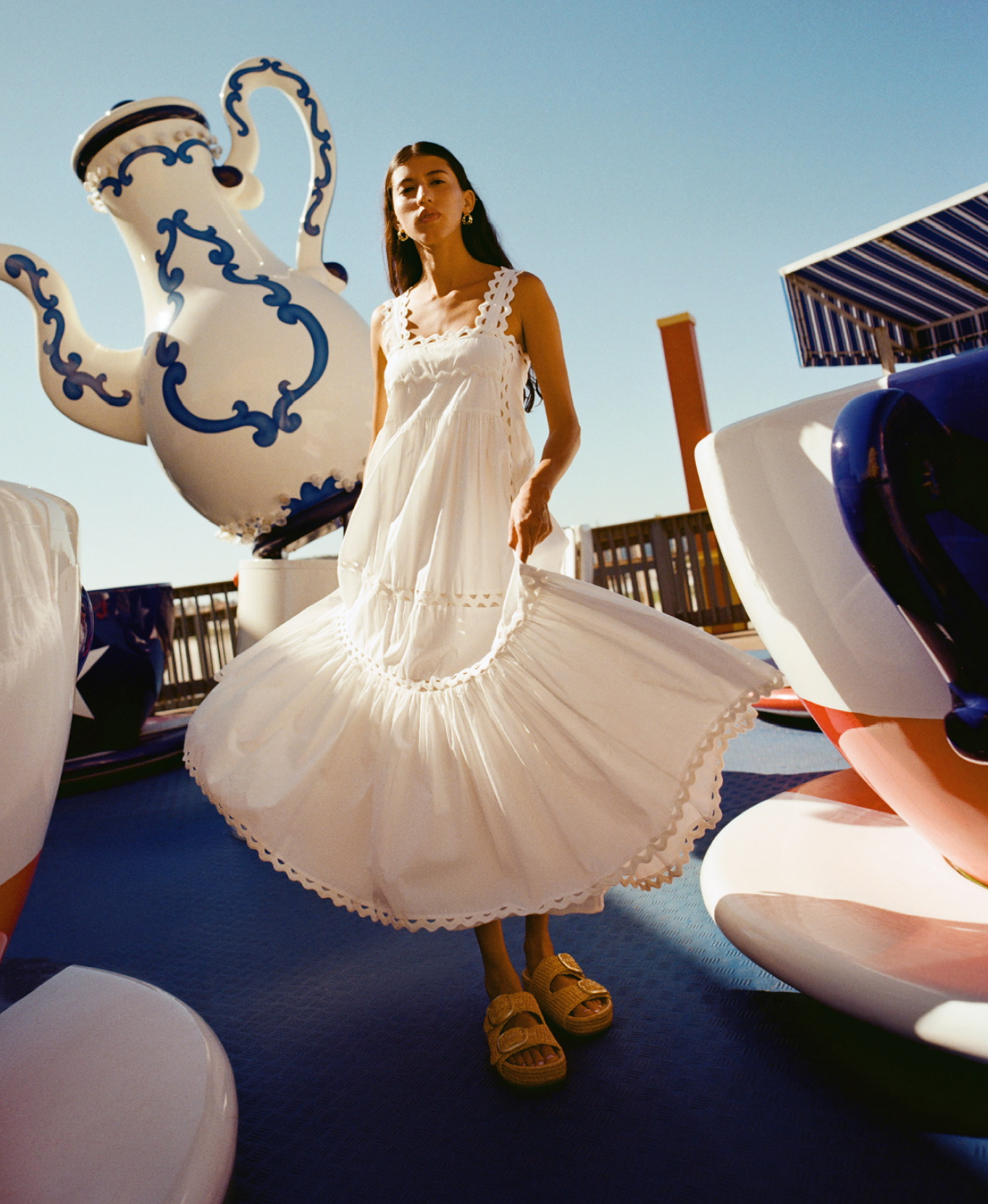 A model stands in front of a teapot statue wearing a white poplin dress and double banded flat sandals.