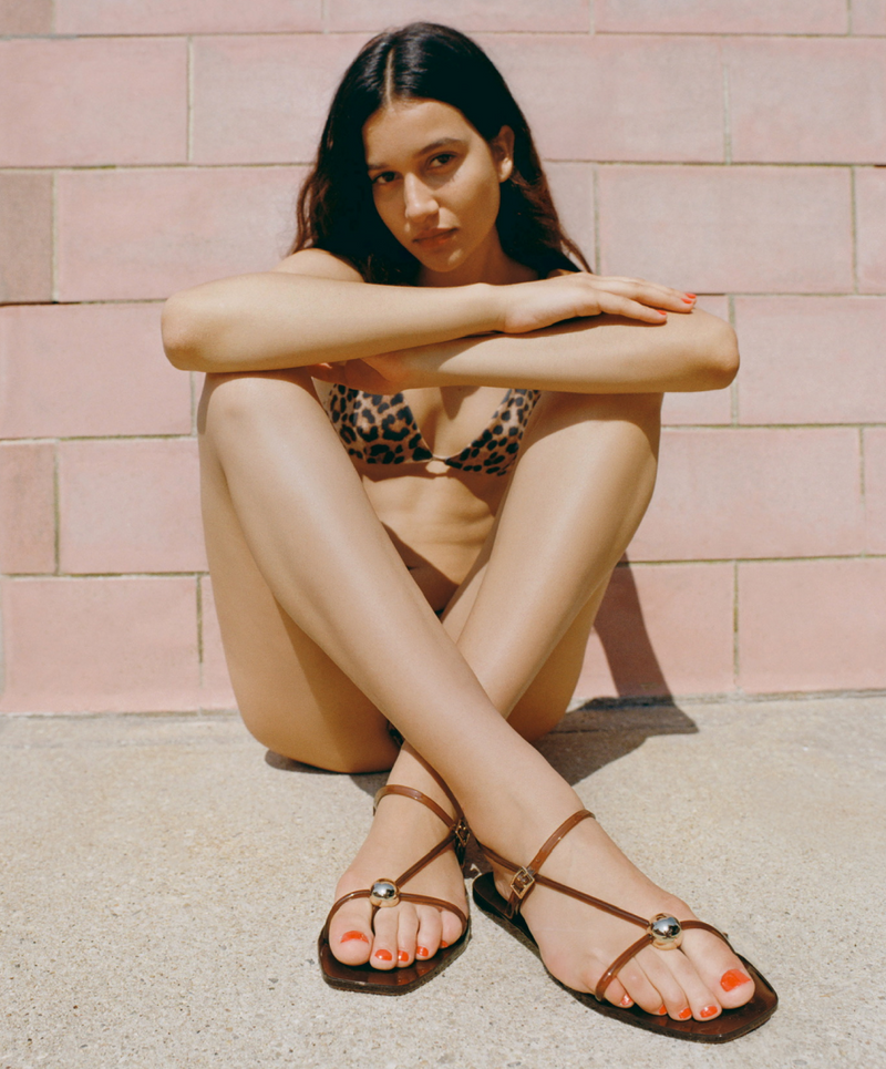 A model sits against a pink brick wall wearing a leopard bikini and brown jelly sandals.