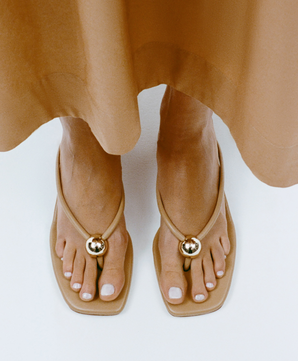 An overhead view of a model wearing tan strappy sandals with a gold ball near the toes and a light brown skirt.