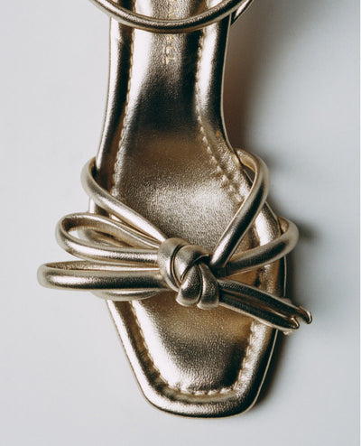 An overhead view of a champagne sandal with a knot detail on a light grey background