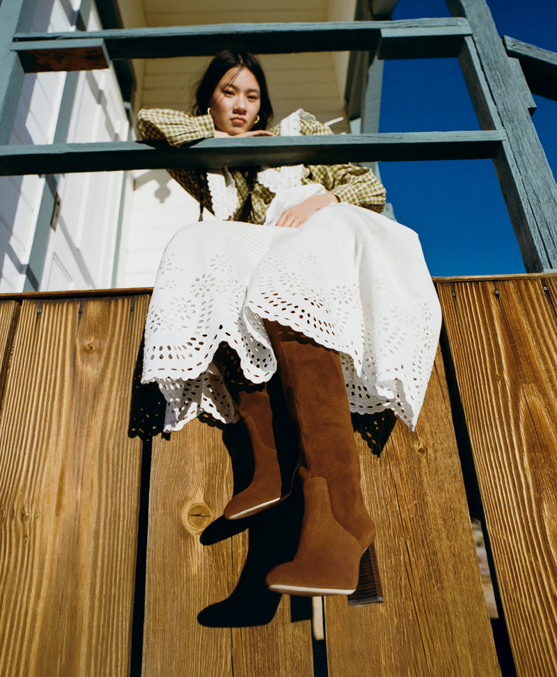 Woman sitting on a wooden railing wearing a white dress and brown suede tall boots.