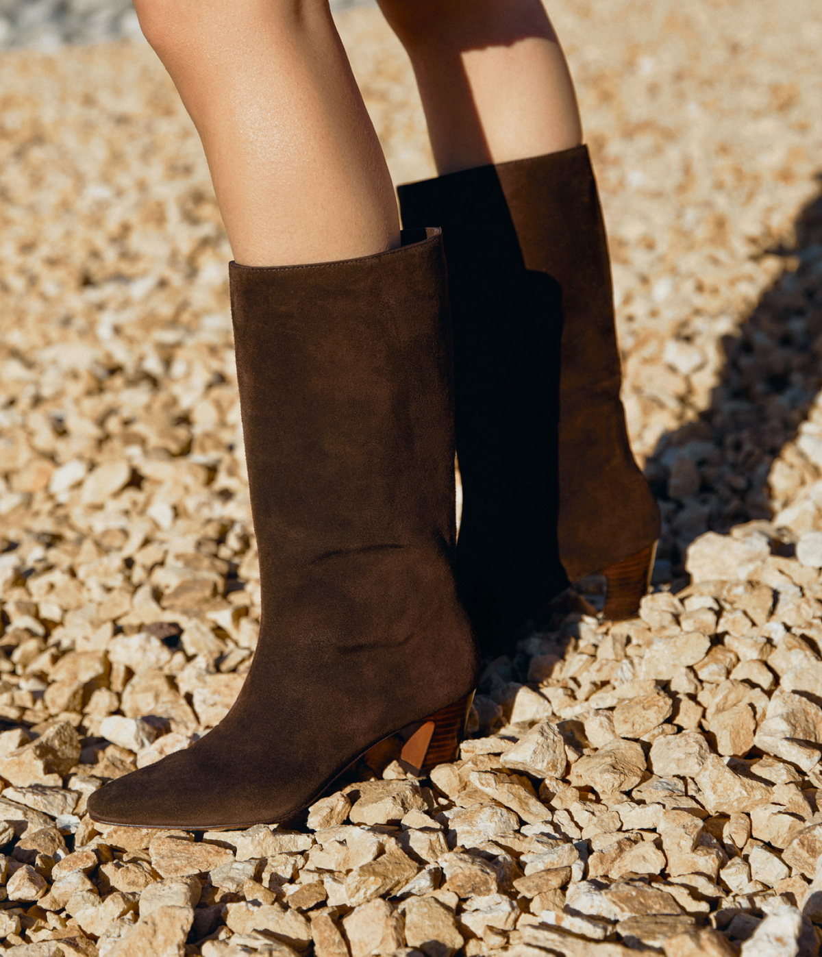 A close up of a pair of brown suede tall boots on a rocky surface.