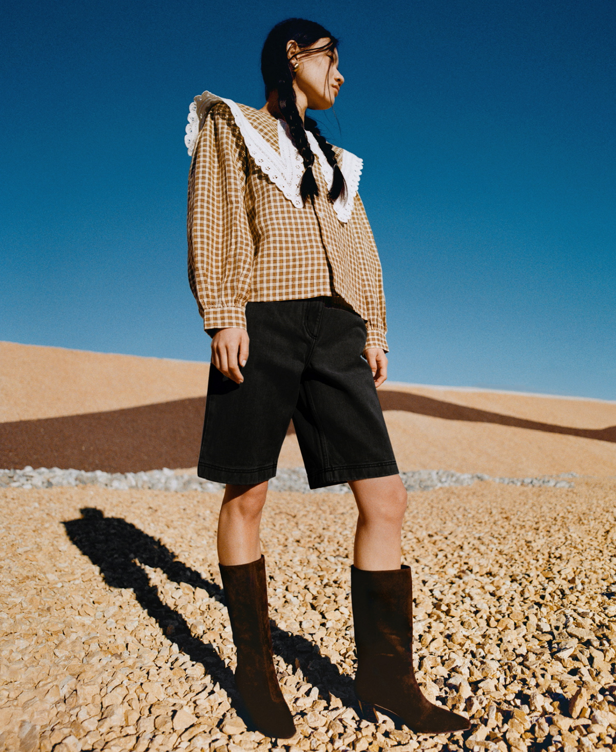 A model stands on a rocky surface wearing black shorts, a brown plaid top with lace accents, and a pair of brown suede boots.