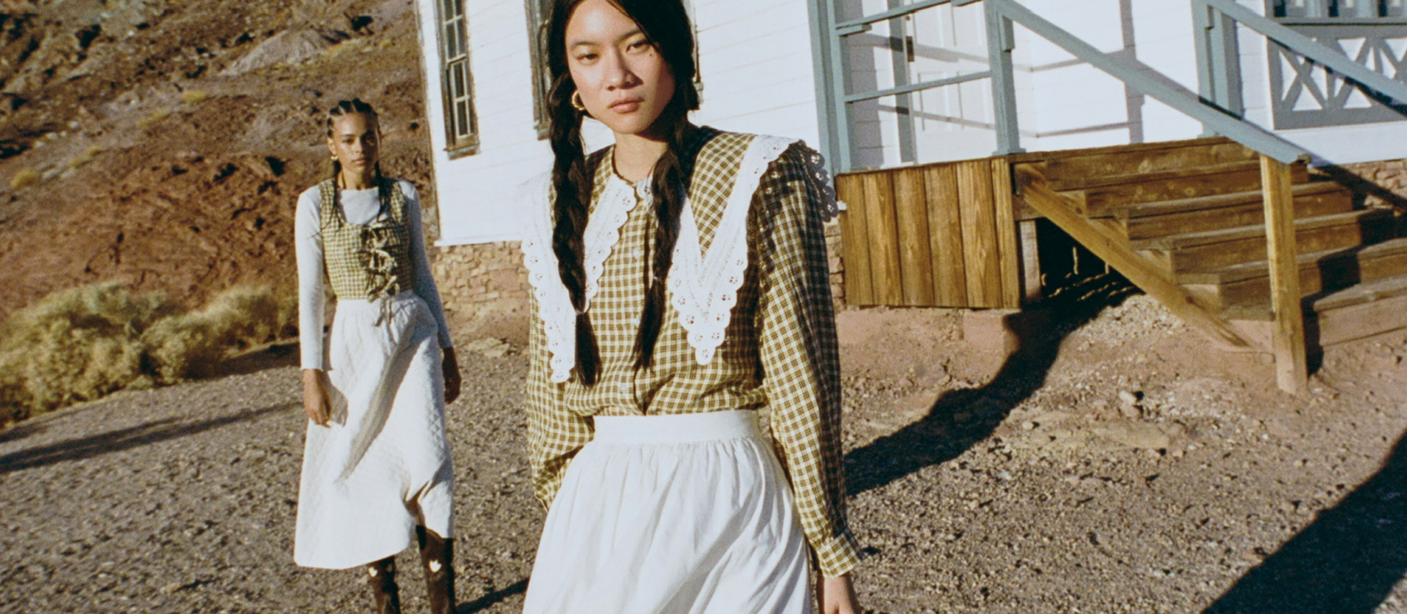 Two women in plaid tops and white skirts standing in a desert-like setting with house in the background.