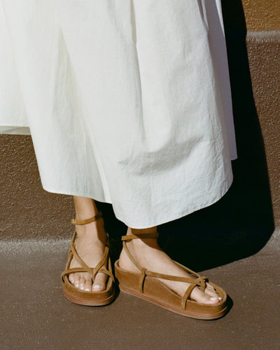 A close up of a model wearing brown suede sandals and a white skirt.