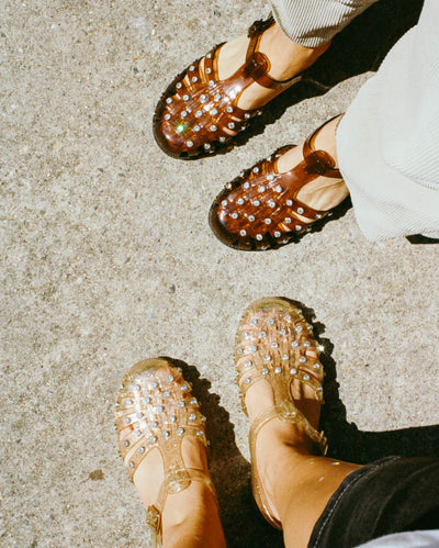 An overhead view of two people standing on a sidewalk wearing jelly sandals, one in brown and one in champagne.