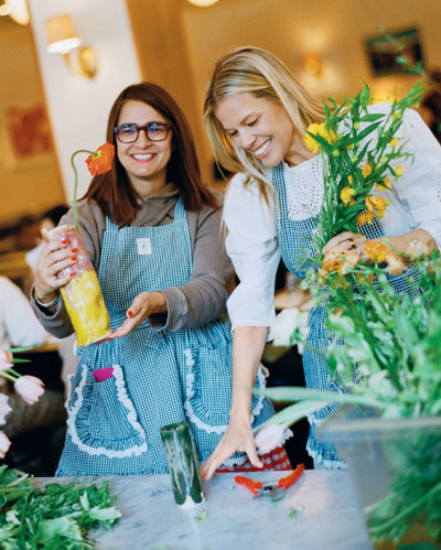 Jessie arranges flowers wearing a white dress and a green gingham apron standing next to a woman wearing the same apron.