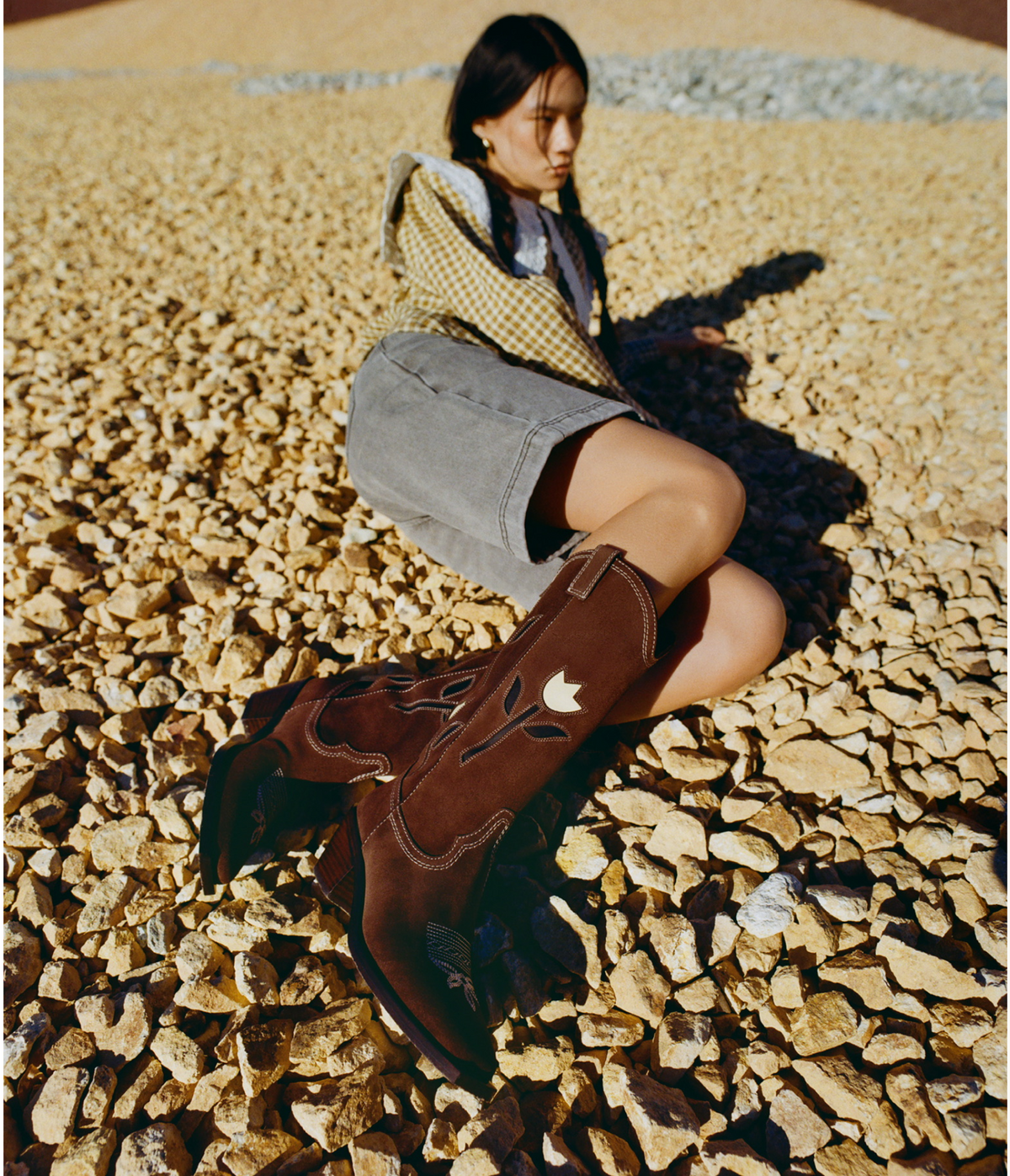 A model lies on her side wearing a pair of brown suede cowboy boots sitting on a rocky pathway.