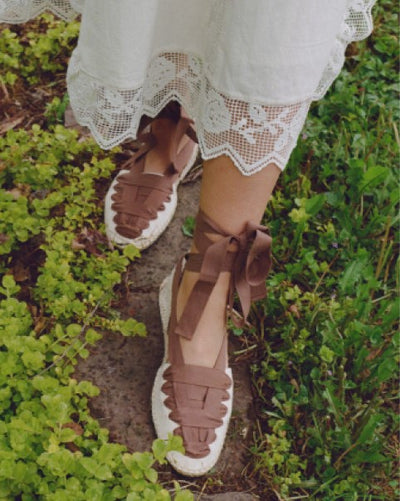 An overhead shot of a model wearing a white lace dress and a pair of brown lace up espadrilles and walking through a green path.