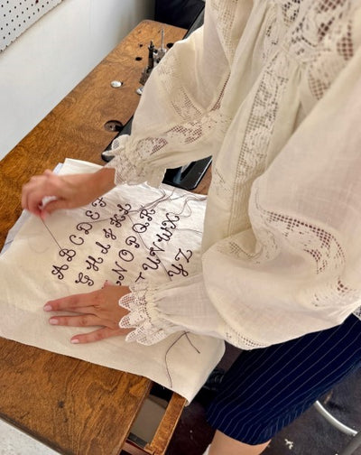Person wearing a white lace blouse working on a sewing project with letters on fabric.