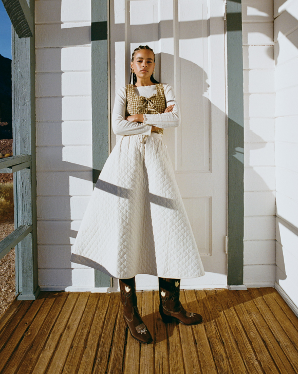 A model leans against a white door wearing a long white skirt with a patterned vest standing on a wooden deck.