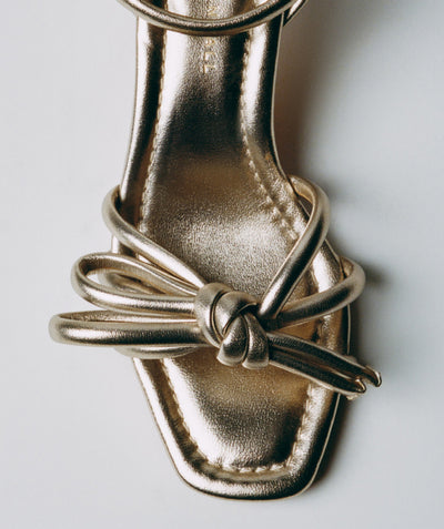 A close up overhead view of a gold sandal with a bow detail on a light grey background.