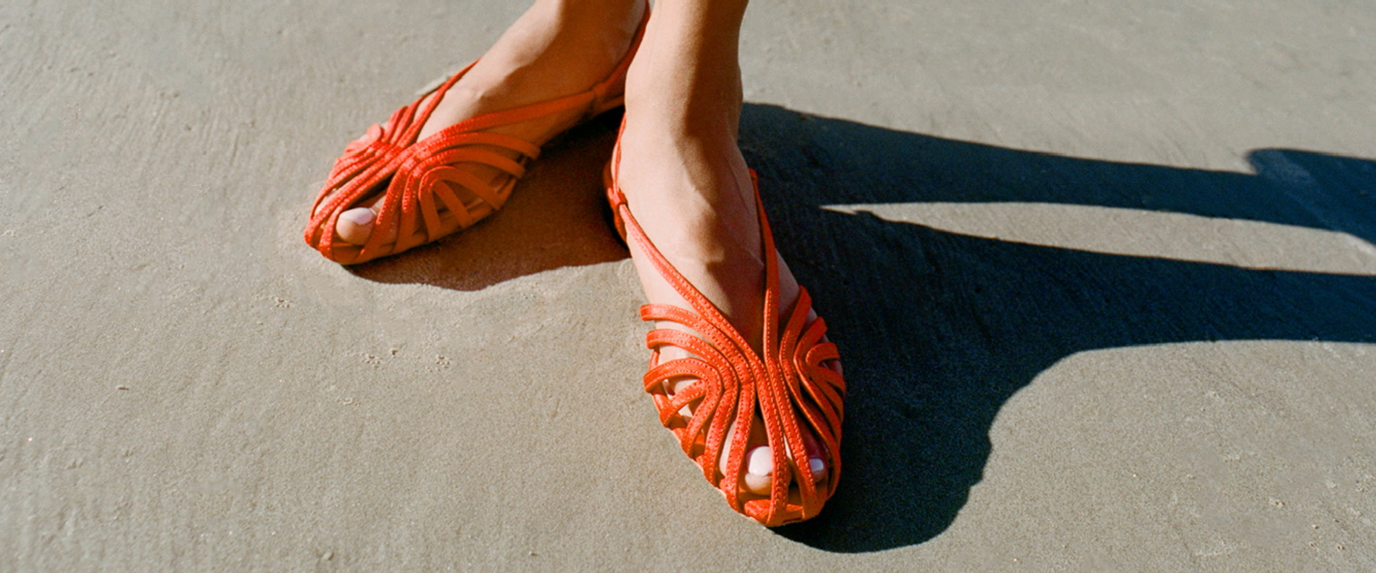 A close up of a model standing on sand wearing red strappy sandals.