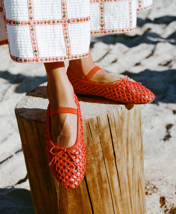 A model stands on a wood pilar wearing red crochet ballet flats and a red and white checkered skirt.