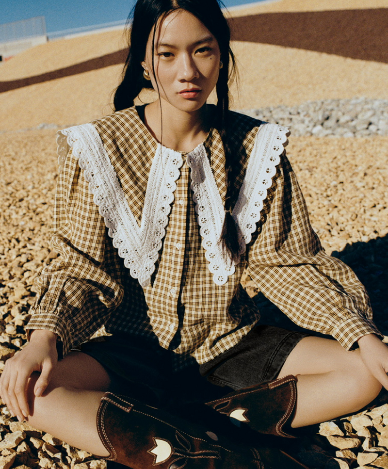 Woman sitting on a rocky surface wearing a checkered blouse with white lace details.