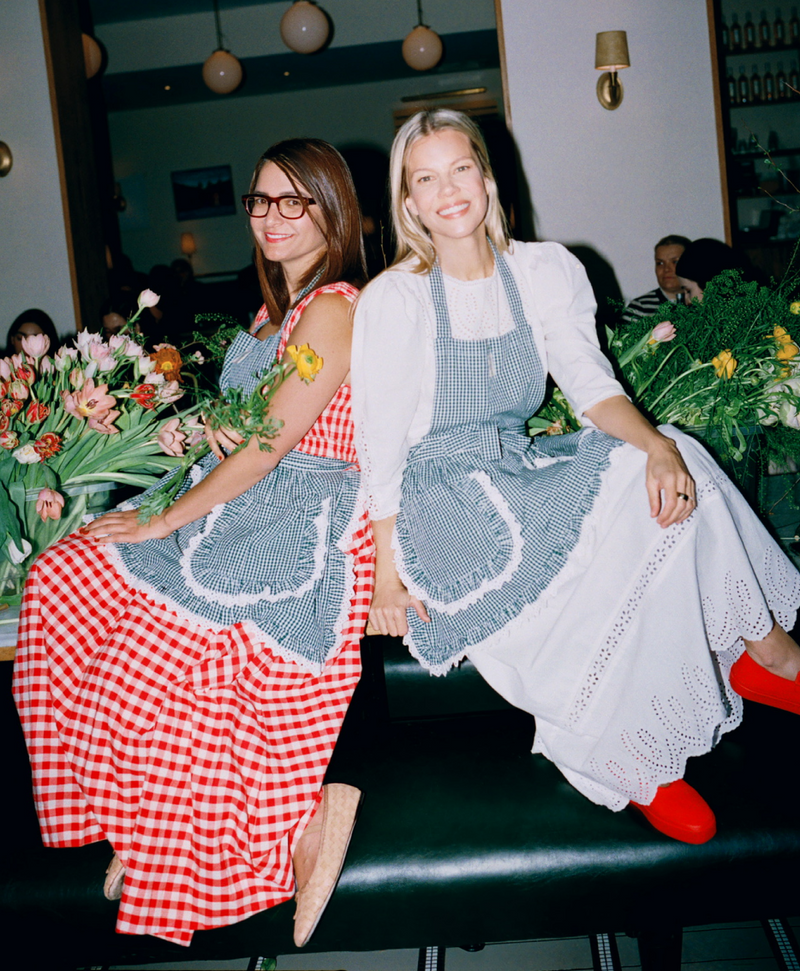 Jessie sits beside two arrangements of flowers and a woman wearing a red gingham dress. Both are wearing green gingham aprons.