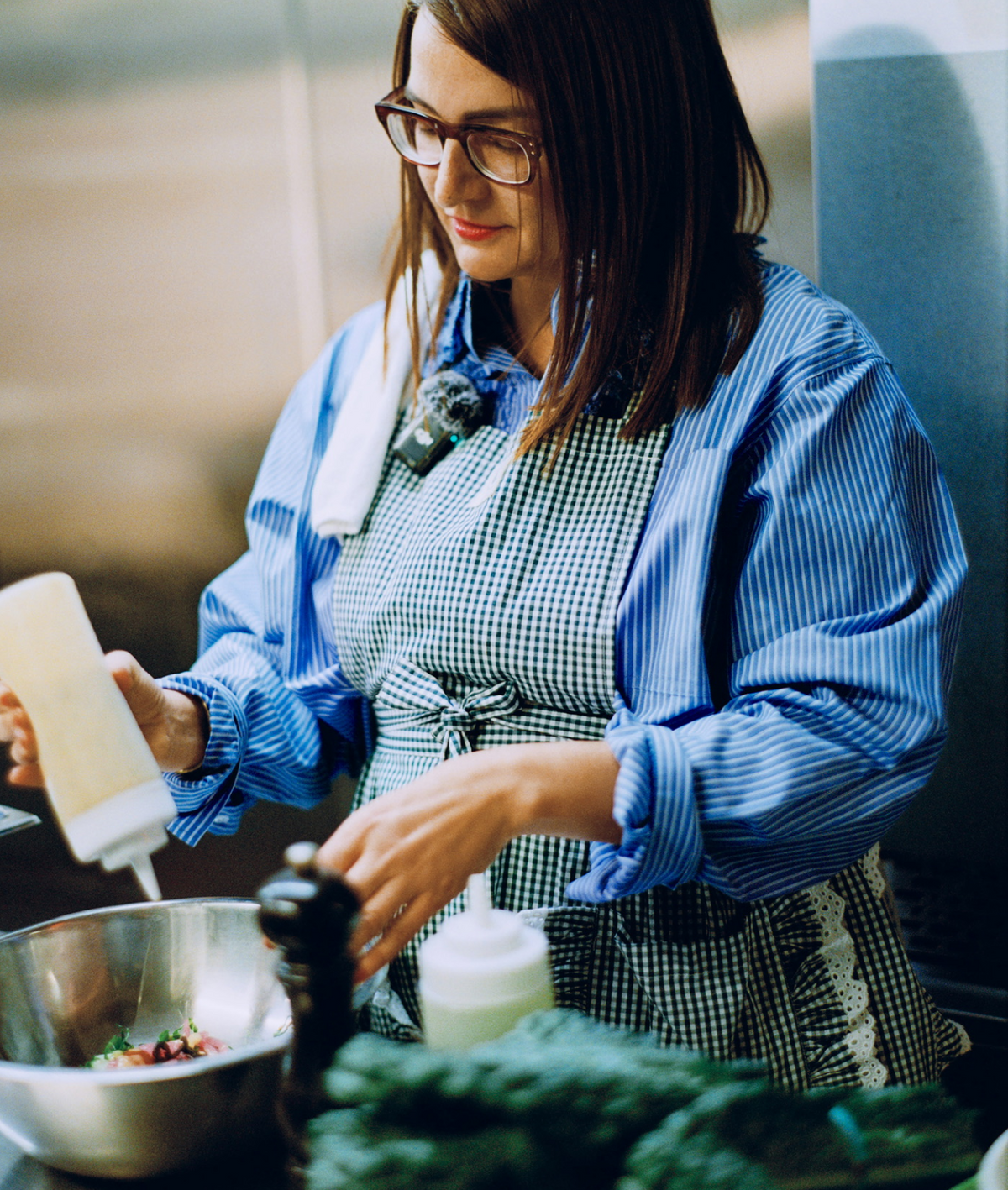 A chef adds dressing to a bowl while wearing a blue and white striped top and a green gingham apron.
